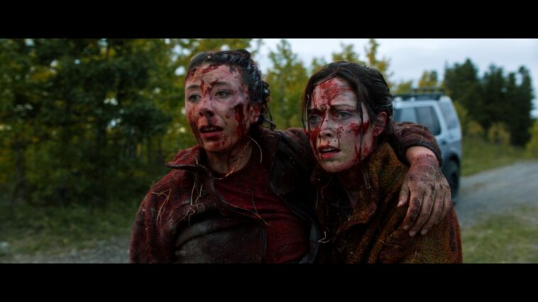 Two bloodied and exhausted people embrace while staring off into the distance.