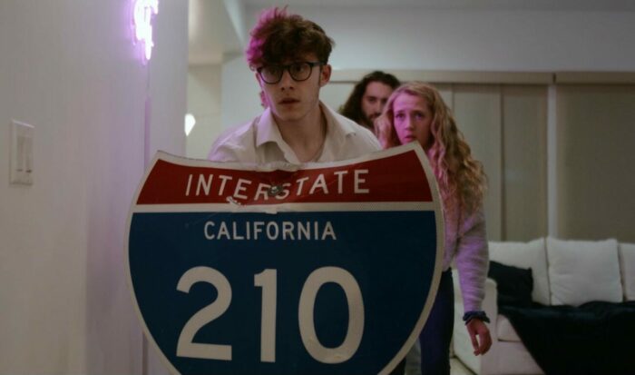 a group of college-aged kids hide behind a shield made out of a California Interstate 210 sign.