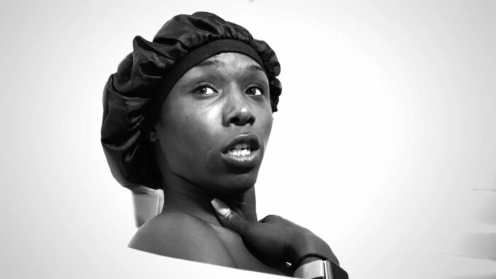 A trans woman sits in her bathtub in a showercap, with her hand on her chest