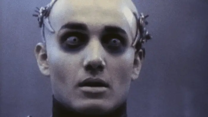 A bald, pale man with a metallic headband, makeup around his eye sockets, and light blue irises stares into the camera