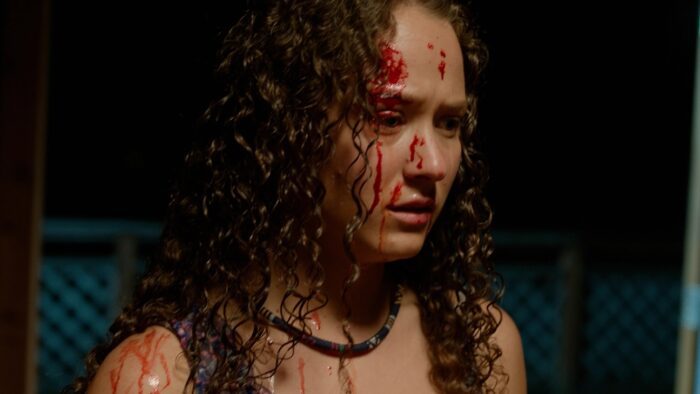 a woman stands shocked, her face is half-covered in blood