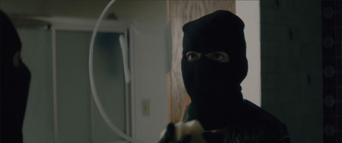 A person in a ski mask looking in a mirror with a happy face drawn on it in American Meltdown