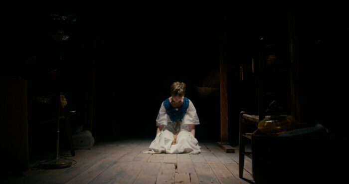 Anya is seen kneeling in a white dress under a spotlight in Mind Body Spirit