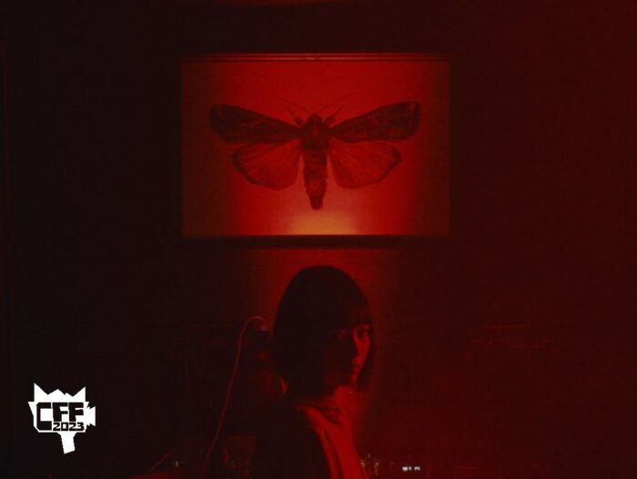 A moth is seen in a frame above Miyabi's head in a room lit by red lights