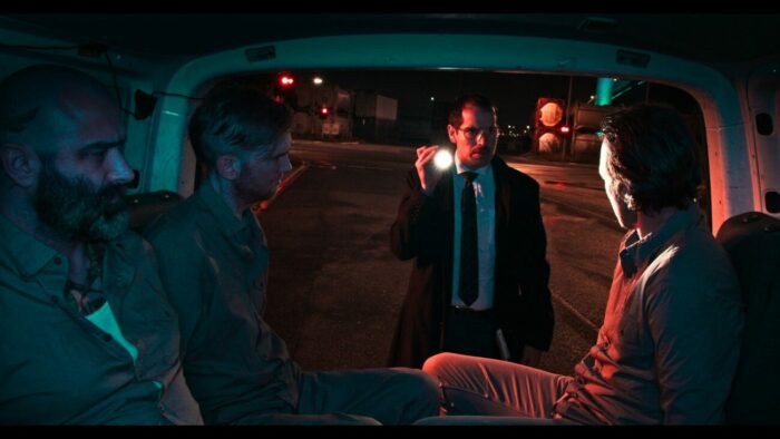 A man standing outside of a van shines a light on a man handcuffed inside while others sit around him in Subject.