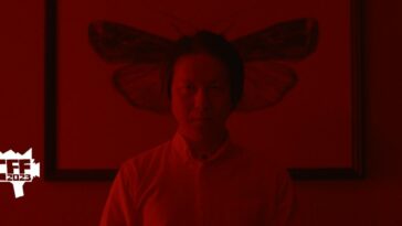 Oka stands in front of a photo of a moth in a red room in New Religion