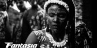 A black and white photo of a woman wearing white seashells in her hair and around her neck and illuminating face paint in Mami Wata