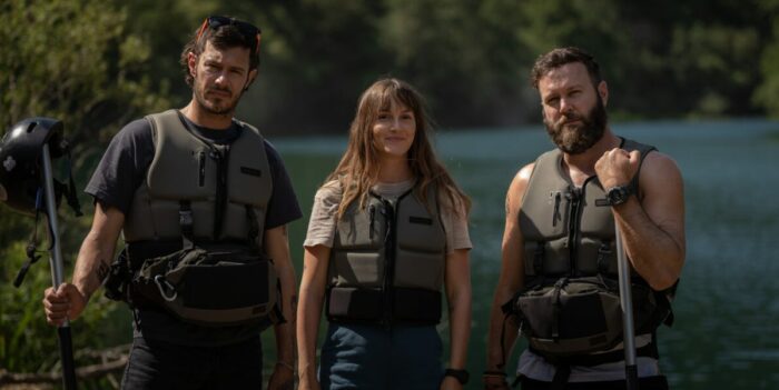Adam Brody, Leighton Meester, and Taran Killam stand together in life preservers on the river bank in River Wild.