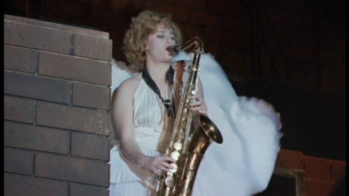 Betty is dressed in a flowing dress, blowing in the wind, as she plays the saxophone