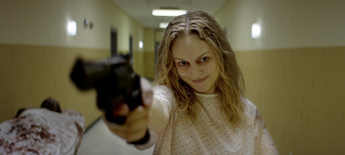 A woman wearing a hospital gown, with matted hair and red around her eyes smiles as she holds a gun toward the camera in Suitable Flesh