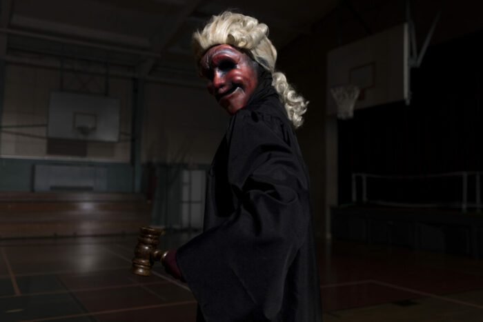 A person wearing a red mask and a colonial wig holds a gavel and looks over their sholder threateningly in Founder's Day