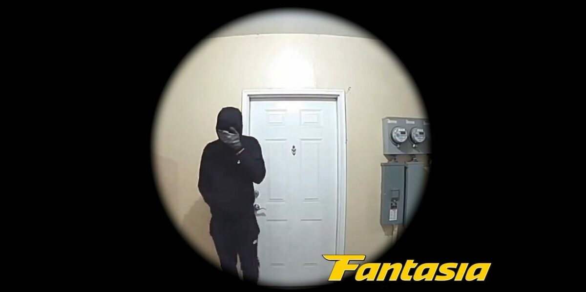 A masked man holds his hand over his face standing with his back to a white door while caught on a doorbell camera in Home Invasion