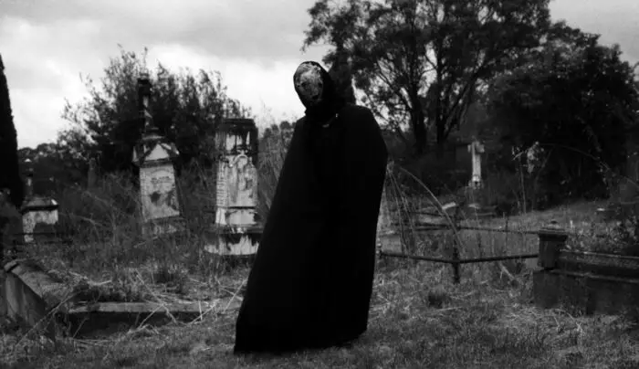 A black cloaked figure stands in the middle of a cemetery. 