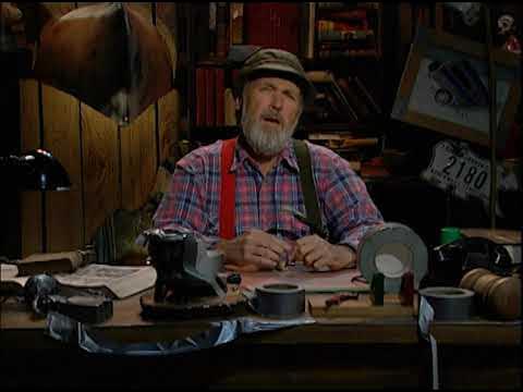 A middle aged, beared white man dressed in flannel and red-green suspenders sits at a desk, covered in various tools and knickknacks.