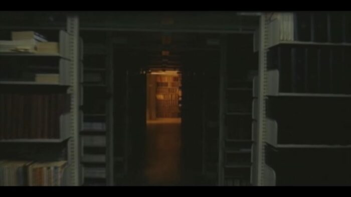 Long shot of the book stacks, with a hallway light shining at the end of the hallway