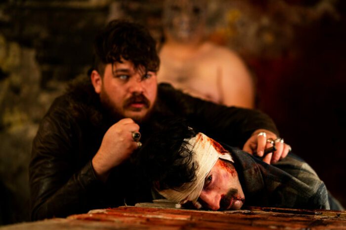 a man with one dead eye holds another man down on a table. The other man's head is wrapped in gauze and blood is seeping through