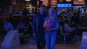 A man stands beside a blond haired woman holding a bowling ball on a bowling alley lane in Saturn Bowling