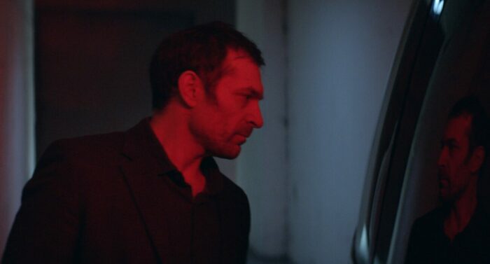 A man with a red light glowing on his face sees his reflection in a car window