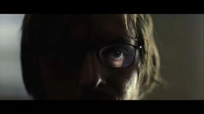 Doug looks through the door, riddled with bullet holes. The light from the outside shines through the bullet hole and over his eye