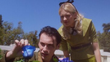 a woman dressed in a conservative Olive dress looks over the shoulder of a man touching a blue rose on a bush