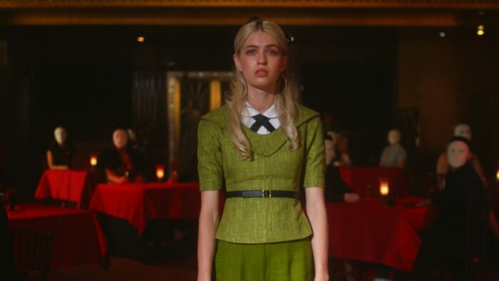 a woman in an olive dress stands mesmerised in a restaurant filled with red tables and masked patrons in The Blue Rose