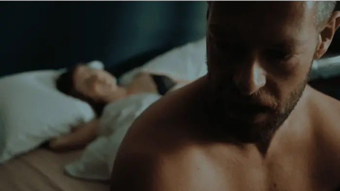A shirtless man sits up in bed while a woman lays under the covers behind him