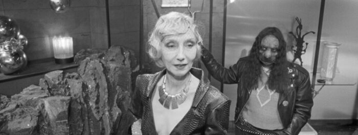 A glamorous older women stands with a woman with the head of a dog leaning on the chair behind her.