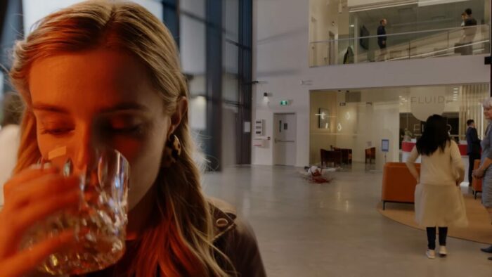 Angela sips a glass of whiskey as a body falls flat to the ground behind her