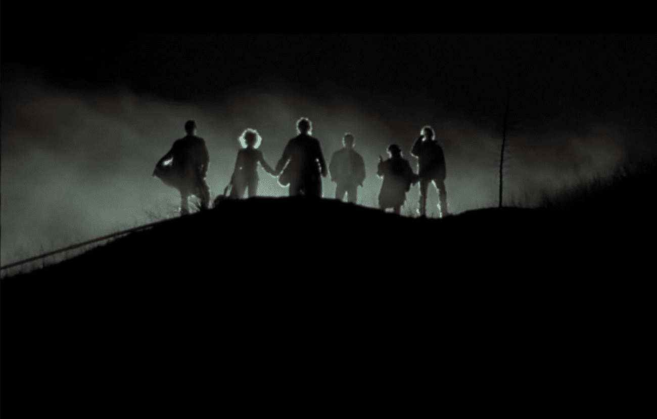 The image depicts a silhouette of six individuals standing on a hilltop against a dark, clouded sky. The lighting from behind creates a dramatic backlight effect, highlighting the contours of their figures. This is a still from the movie "Near Dark" (1987). The group appears to be standing close together, some holding hands, which conveys a sense of unity or alliance among the characters. The atmosphere is eerie and suggests a tense or suspenseful moment in the film.