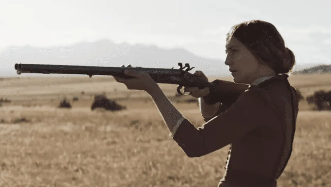 A woman in period clothing aiming a rifle in a vast, open field with mountains in the background.