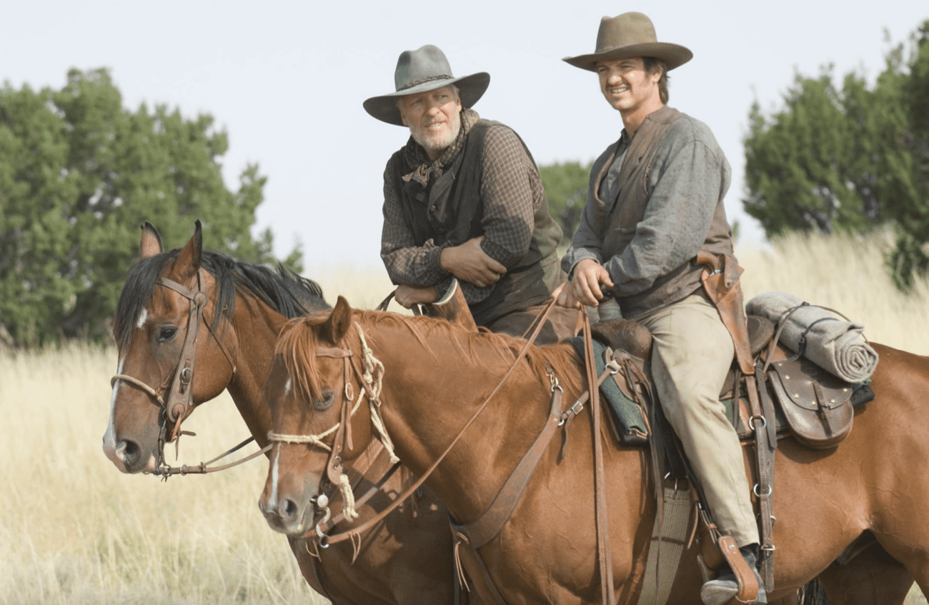 Two men on horseback in a grassy plain, with trees in the background. The man on the left is wearing a wide-brimmed hat, a vest, and a bandana around his neck, portraying a rugged look. The man on the right, also in a wide-brimmed hat, has a more youthful appearance and is dressed in a long-sleeved, dirt-stained shirt, suggesting a setting that could be from a historical or western-themed film. Both men are smiling and appear to be engaged in a light-hearted moment during an outdoor scene.