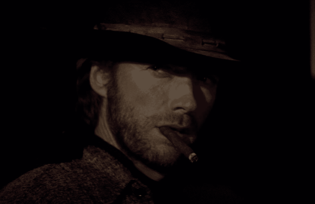 A man with a rugged look, wearing a dark cowboy hat, is seen in a dimly lit setting. He has stubble on his face and is staring intently to the side. A cigarette is placed in the corner of his mouth, emitting a thin trail of smoke. The overall atmosphere is moody and reminiscent of classic Western films.