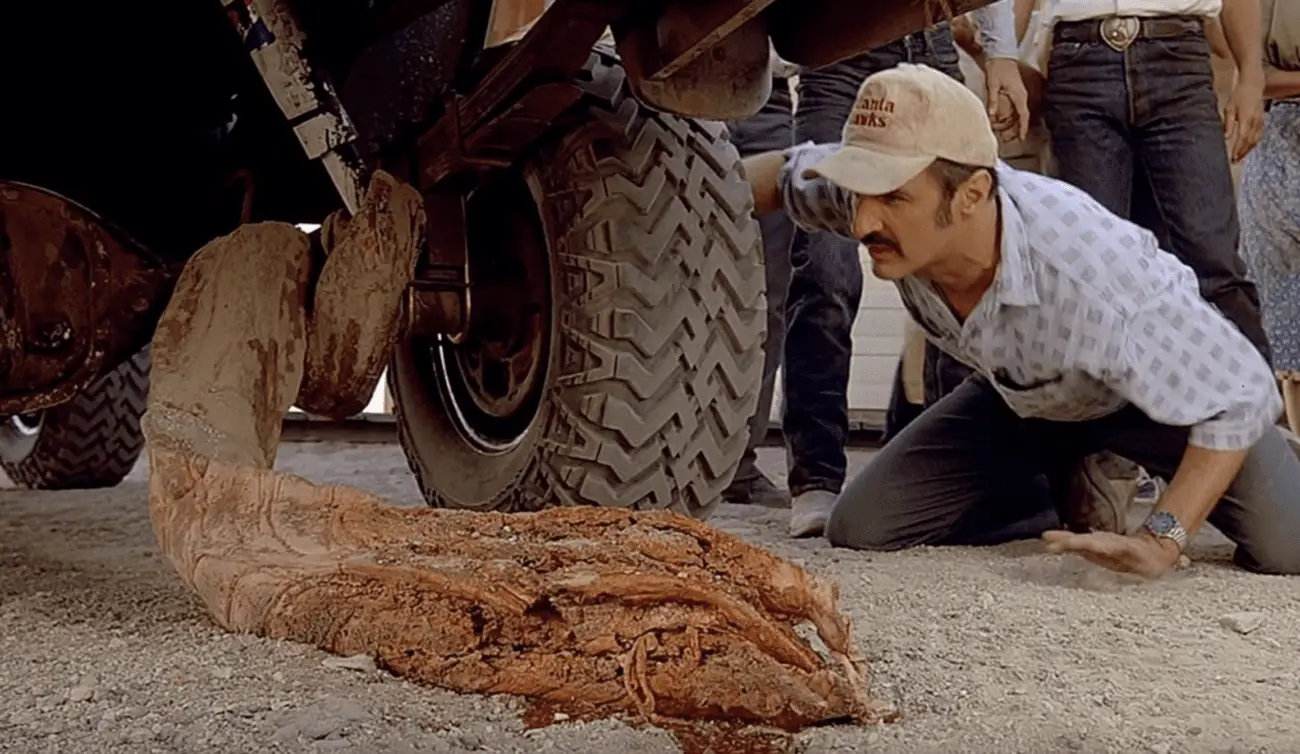 A man is crouched on a dusty ground next to a large worm-like creature, examining it closely. He is wearing a cap that says "Atlanta Hawks," a light-colored, patterned shirt, and dark pants. Behind him, various onlookers watch intently. The worm creature appears to be dead or incapacitated, and its body is partially under the wheel of a large vehicle, suggesting a scene from a movie involving monstrous creatures. This scene is set in a dry, outdoor location, likely representing a rural or desert area.