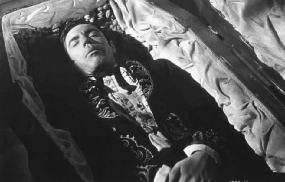 A man in an embroidered outfit is lying with closed eyes in a coffin, from the movie "Curse of the Undead" (1959).