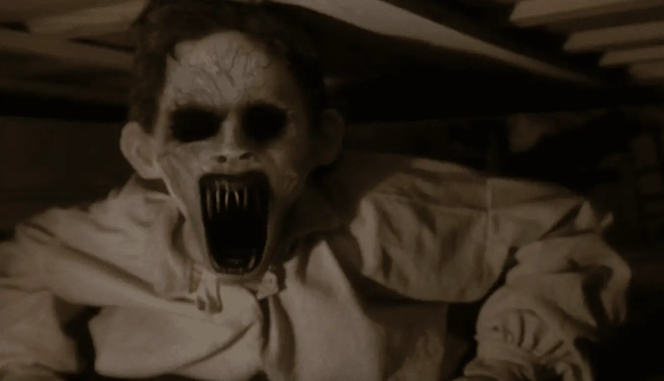 A person with pale, sickly skin and dark circles under their eyes, mouth agape showing elongated, sharp teeth, giving them a ghoulish appearance. They're dressed in a simple, beige, old-fashioned shirt and appear to be in a dimly lit, cramped space with wooden beams, suggesting a horror scene from a movie.