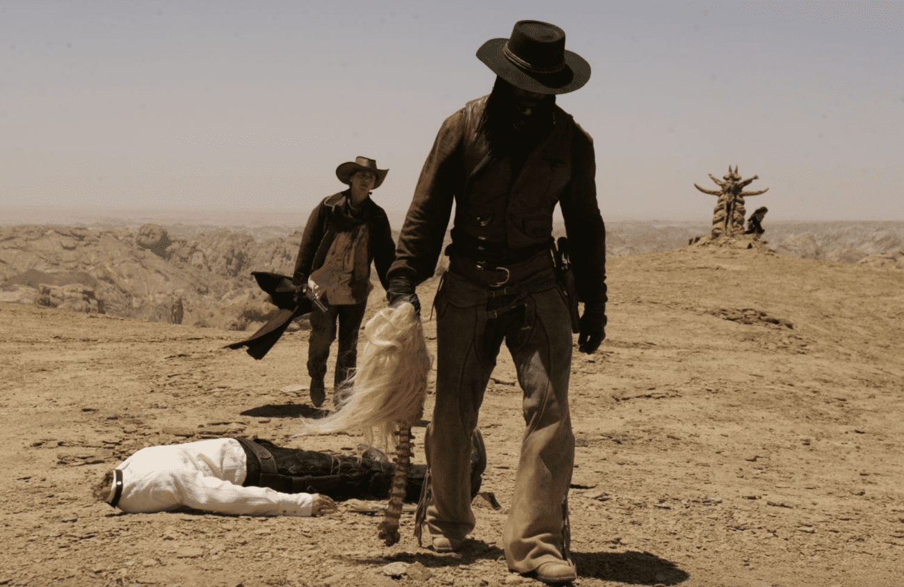 A scene from the film 'Gallowwalkers' featuring two characters in a desert landscape. The character in the foreground is dressed in a dark cowboy hat, a maroon shirt, and leather pants, holding a blonde wig in one hand. In the background stands another character, appearing dusty and worn, holding a shovel over his shoulder. The ground is barren and rocky, with a clear blue sky overhead. In the distance, there is a peculiar wooden structure, adding to the desolate Western atmosphere of the scene.