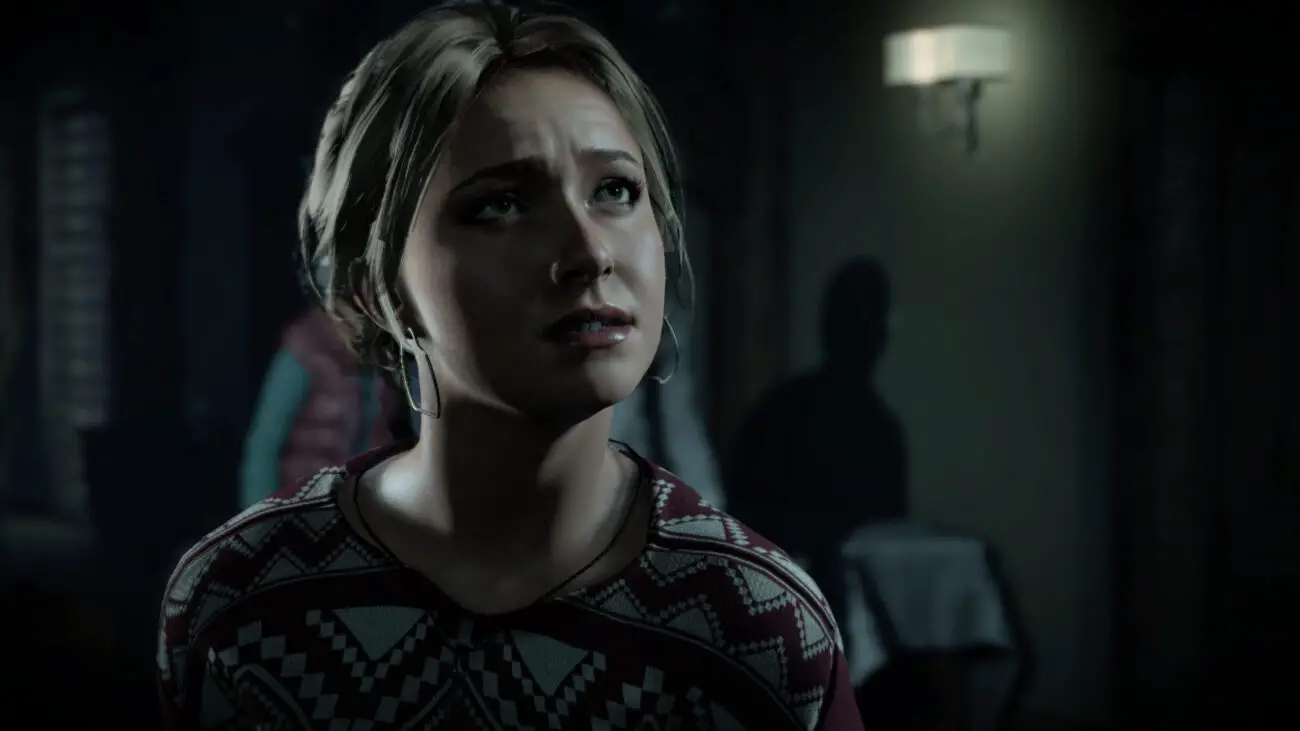 A digital image of a young woman from a video game "Until Dawn". She has blonde hair, hoop earrings, and is wearing a red and white patterned sweater. Her expression is one of worry or fear, as she looks away from the camera with wide eyes and a slightly open mouth. The background is dimly lit and features an out-of-focus silhouette of another character in the shadows, adding to the suspenseful atmosphere of the scene.