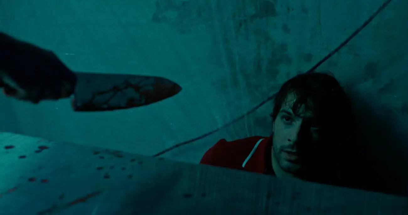 A tense moment from a dark scene where a fearful man is looking up, his face partially obscured by a ledge. Above him, an ominous hand brandishes a bloody knife, suggesting imminent danger or a violent act. The lighting is low and the color palette