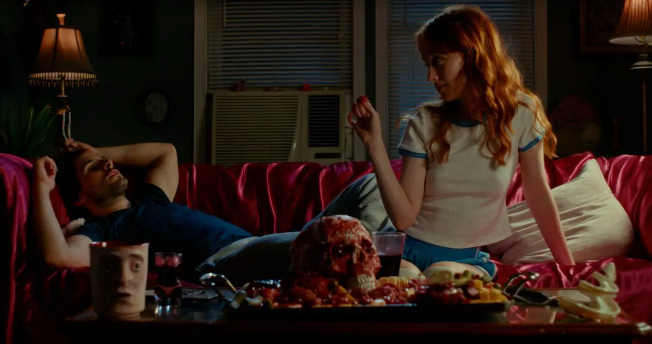 A man lounges on a plush red sofa, appearing lethargic or possibly asleep, with one arm resting on his forehead. A woman sits next to him, her body turned towards him, mid-gesture as if in conversation or making a point. She has long, reddish hair and is dressed in a casual, retro-style outfit. In front of them on a wooden tray is a disconcerting array of items, including a mug with a face design, a glass of red liquid, a skull, and an assortment of foods that seem to include meats and cheeses in a chaotic display. The room is dimly lit, with a window air conditioner in the background and a vintage lamp providing a soft glow to the scene.