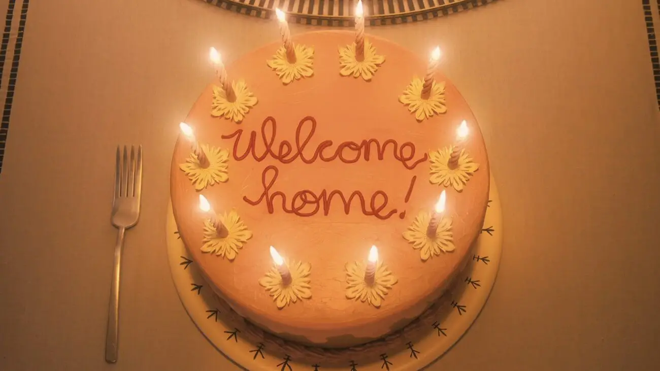 A pink cake with the words "Welcome Home" written in red icing is surrounded by lit candles, placed on a table with a single fork to the left.