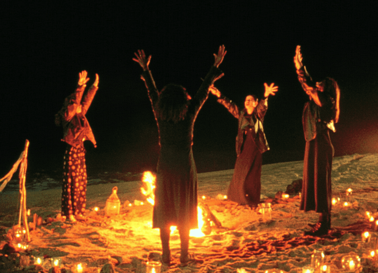 Four young women stand around a bonfire on a beach at night, their arms raised and faces illuminated by the fire's glow. They are engaged in a ritual, surrounded by lit candles and lanterns. The scene exudes an air of supernatural mystery, reflective of the film 'The Craft' (1996).
