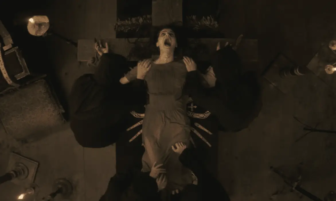 Image shows a woman lying on a large wooden crucifix, dressed in a simple, brown, long-sleeved dress. She is screaming in terror, her mouth wide open and eyes shut tight. Four hooded figures, clad in black robes, are pinning her down, each holding her limbs to the cross. The room is dimly lit by candles, creating a dark and ominous atmosphere. This intense scene is from the 2017 film "The Crucifixion," starring Ada Lupu, depicting the harrowing ordeal of a modern-day exorcism.