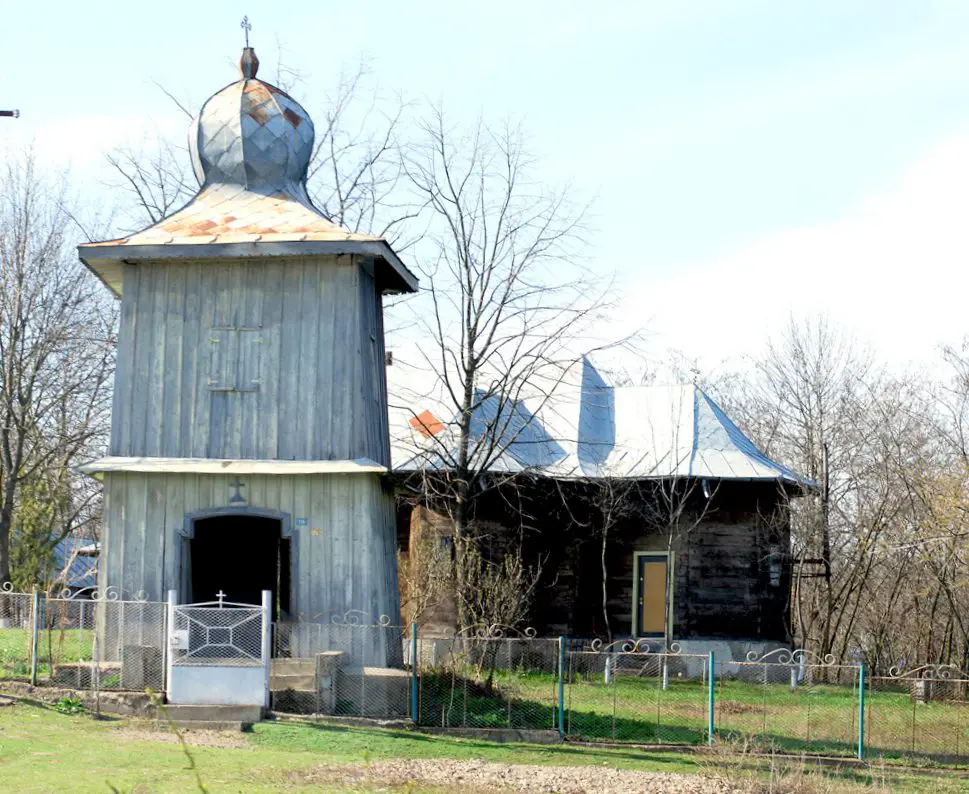 **Image Description:**This image shows the Church of the Holy Governors from Tanacu along with its bell tower, as seen from the south view in April 2010. The church is a rustic wooden structure with a corrugated metal roof, reflecting its historical and traditional architecture. The bell tower, also made of wood with a similarly styled roof, stands prominently in the foreground, showcasing a cross atop its onion-shaped dome. A small gate and a fence encircle the churchyard, and the surrounding trees are bare, indicating early spring. The sky above is clear, contributing to the serene and timeless atmosphere of the scene.