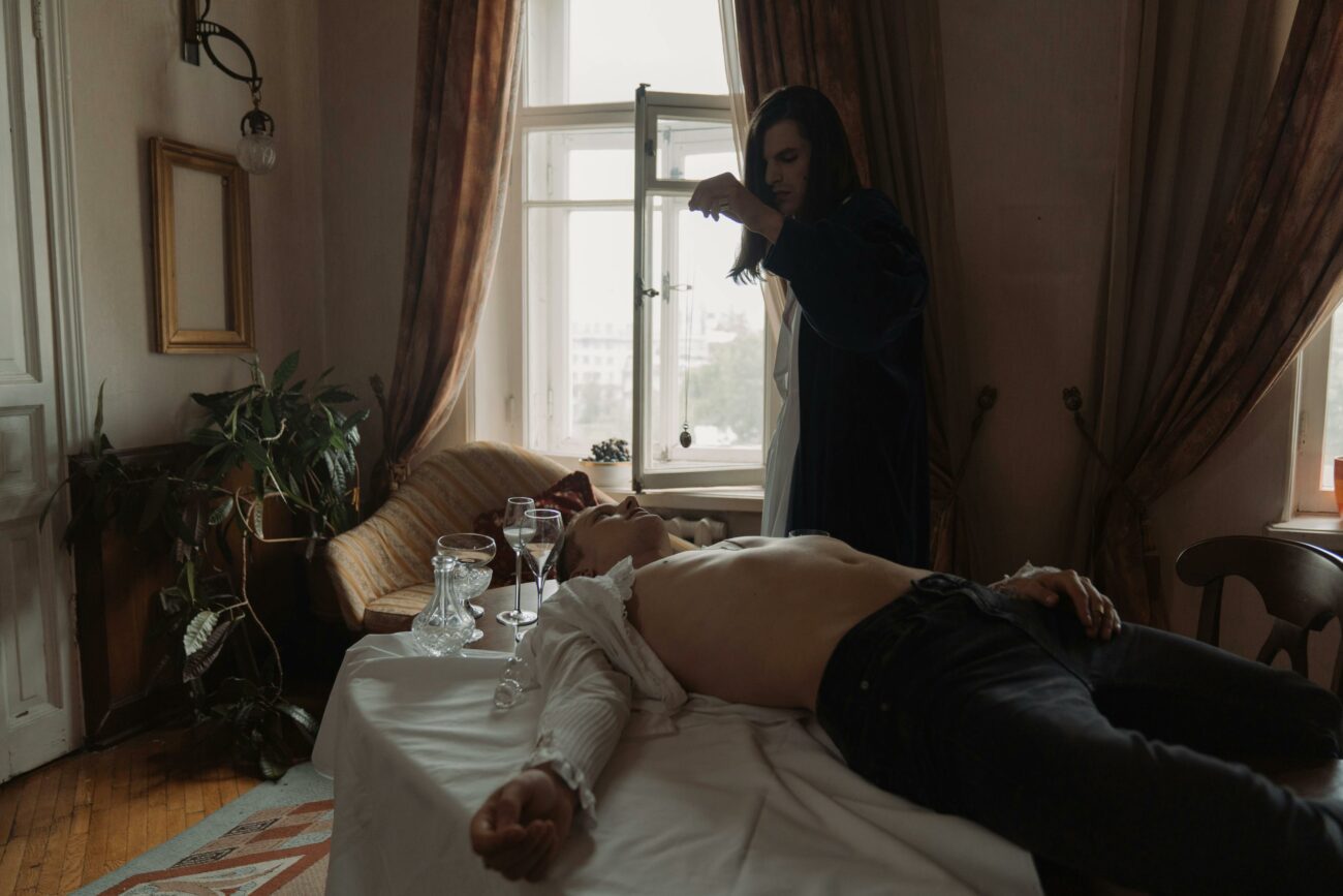 A dramatic scene of a modern-day exorcism. A young man lies unconscious on a table with his shirt partially open, exposing his chest. Another individual, dressed in a dark robe, stands beside him, holding a pendant or amulet over his body. The room is dimly lit, with light filtering through a window behind them, casting a somber atmosphere. The setting includes an old-fashioned interior with heavy drapes, houseplants, and a few crystal glasses on the table, evoking a sense of eerie ritual and intense spiritual confrontation.