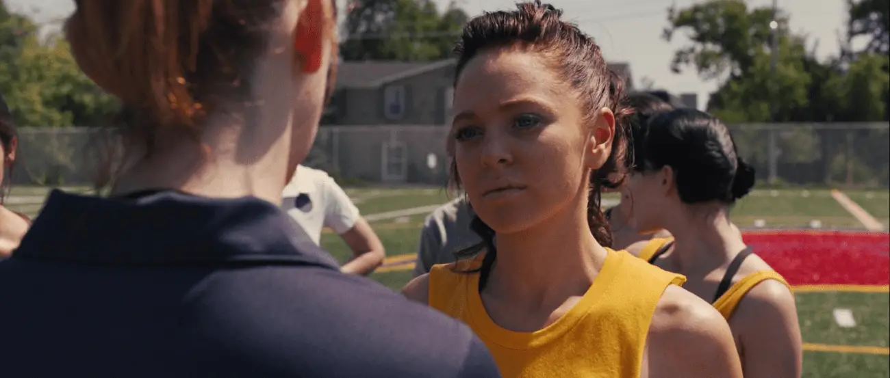 A scene from "Carrie" (2013) with Gabriella Wilde as Sue Snell on a school sports field, staring intensely at another character. The image underscores the tense dynamics among the students.