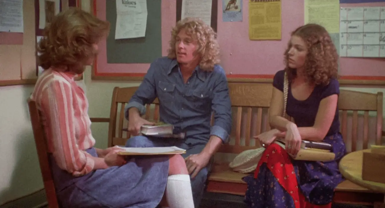 A scene from "Carrie" (1976) featuring actors in a high school setting, dressed in 1970s fashion, discussing school-related matters. The image highlights the period-specific elements of the original film.