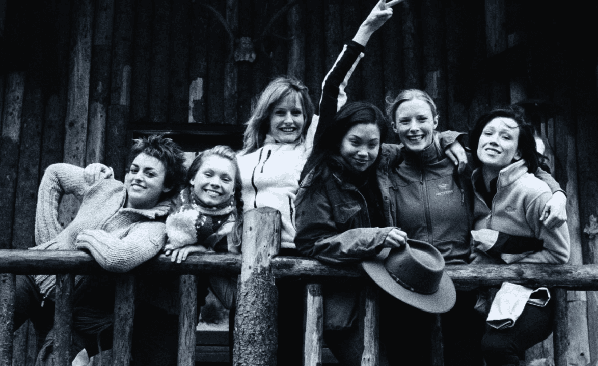 A black-and-white photo showing six women standing on a wooden balcony, smiling and posing together. They are dressed in warm clothing, with some wearing jackets and one holding a hat. The setting appears to be a rustic cabin, suggesting a close-knit group enjoying an outdoor adventure.