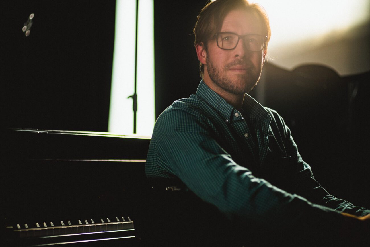 Chris Thomas sitting by a piano, illuminated by soft lighting in a studio setting.