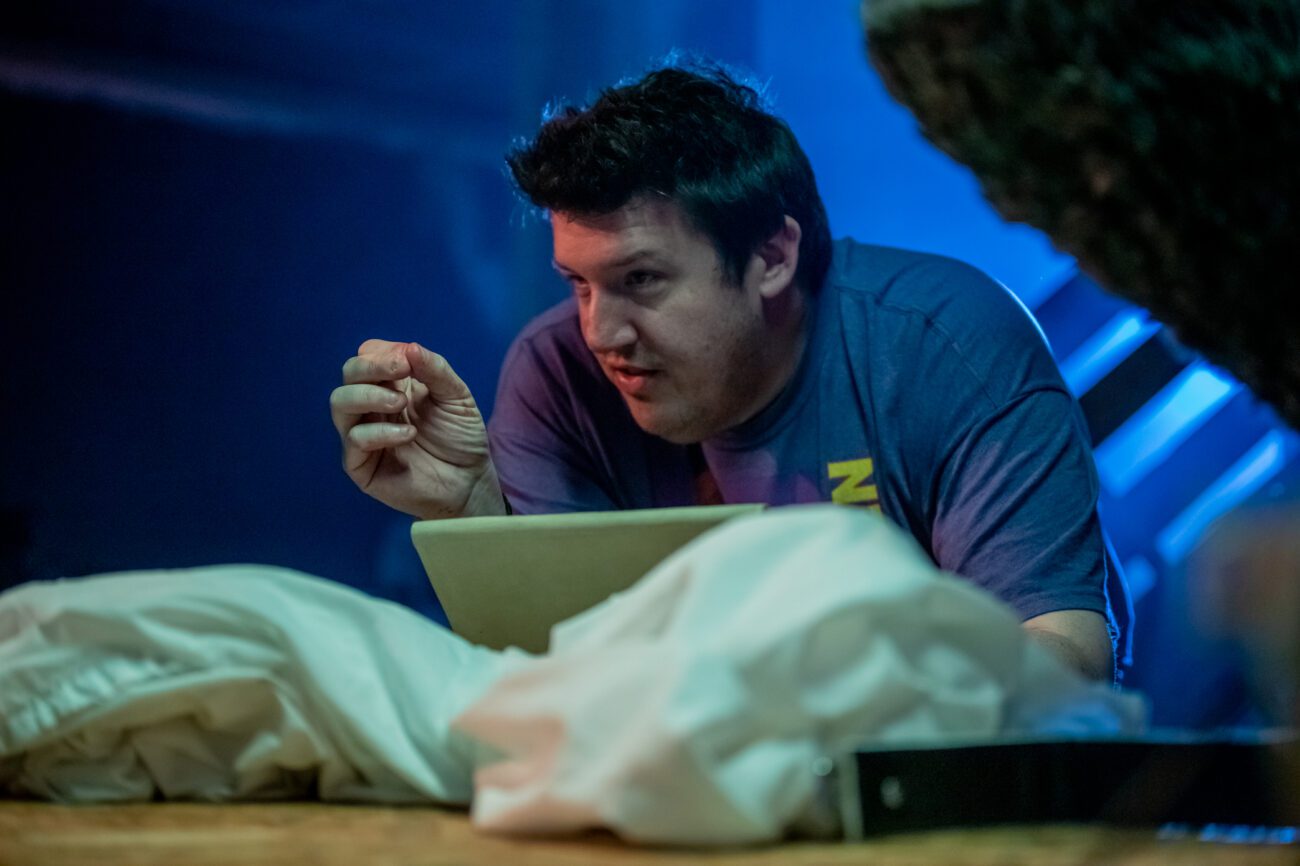 The director kneels near a horror set, gesturing passionately while giving instructions to the cast or crew. The dark setting around him hints at the tension of the scene being filmed.