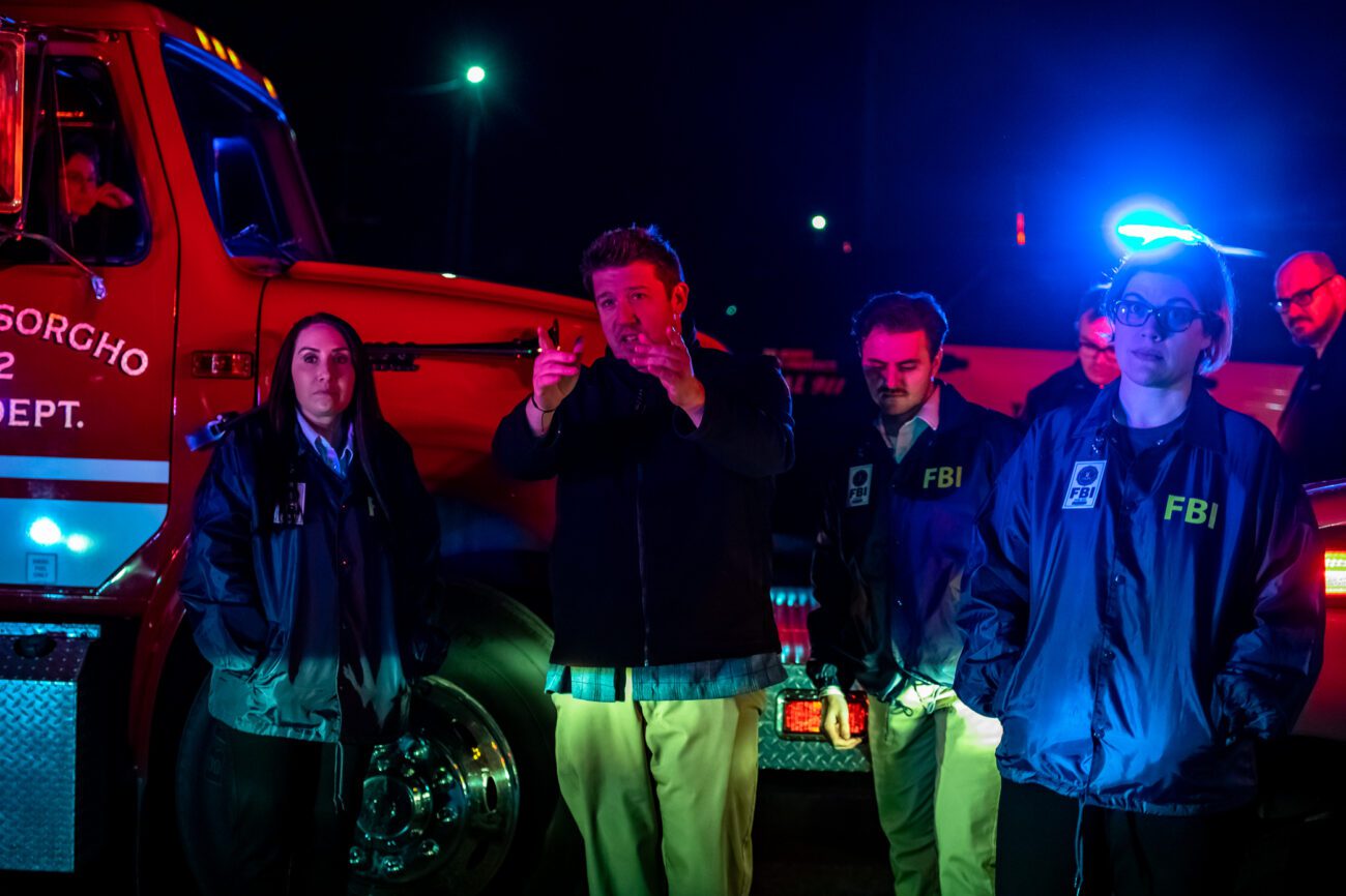 The director, surrounded by actors dressed as FBI agents, gestures while explaining a scene during a night shoot. The scene is lit by the flashing lights of emergency vehicles, adding a tense, cinematic feel.